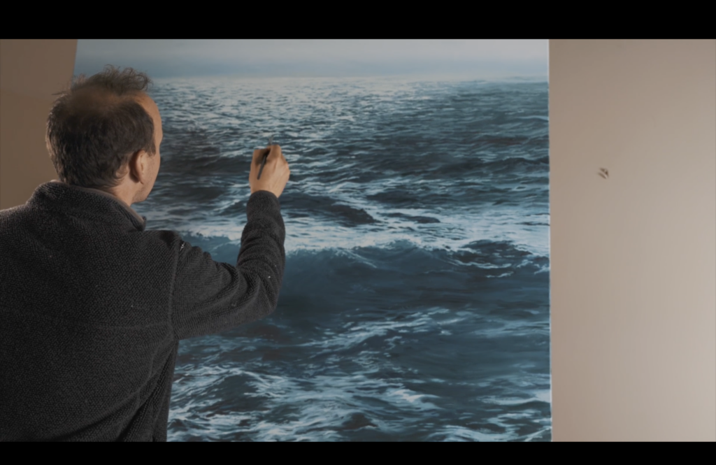 A photographic still from the short film The Artist. The photo shows the artist painting the ocean on a large canvas.
